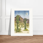 Fine art print by Houston based artist Jenny Wagner, Desert Moon. This watercolor print depicts a desert scene with saguaro cacti and a full moon in the background. Print shown leaning against a neutral wall