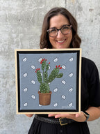 Texas artist Jenny Wagner holding her mixed media original painting, Prickly. This painting features a hand stretched bandana with a potted prickly pear cactus in acrylic paint, with hand beaded glass beads and sequins on top.