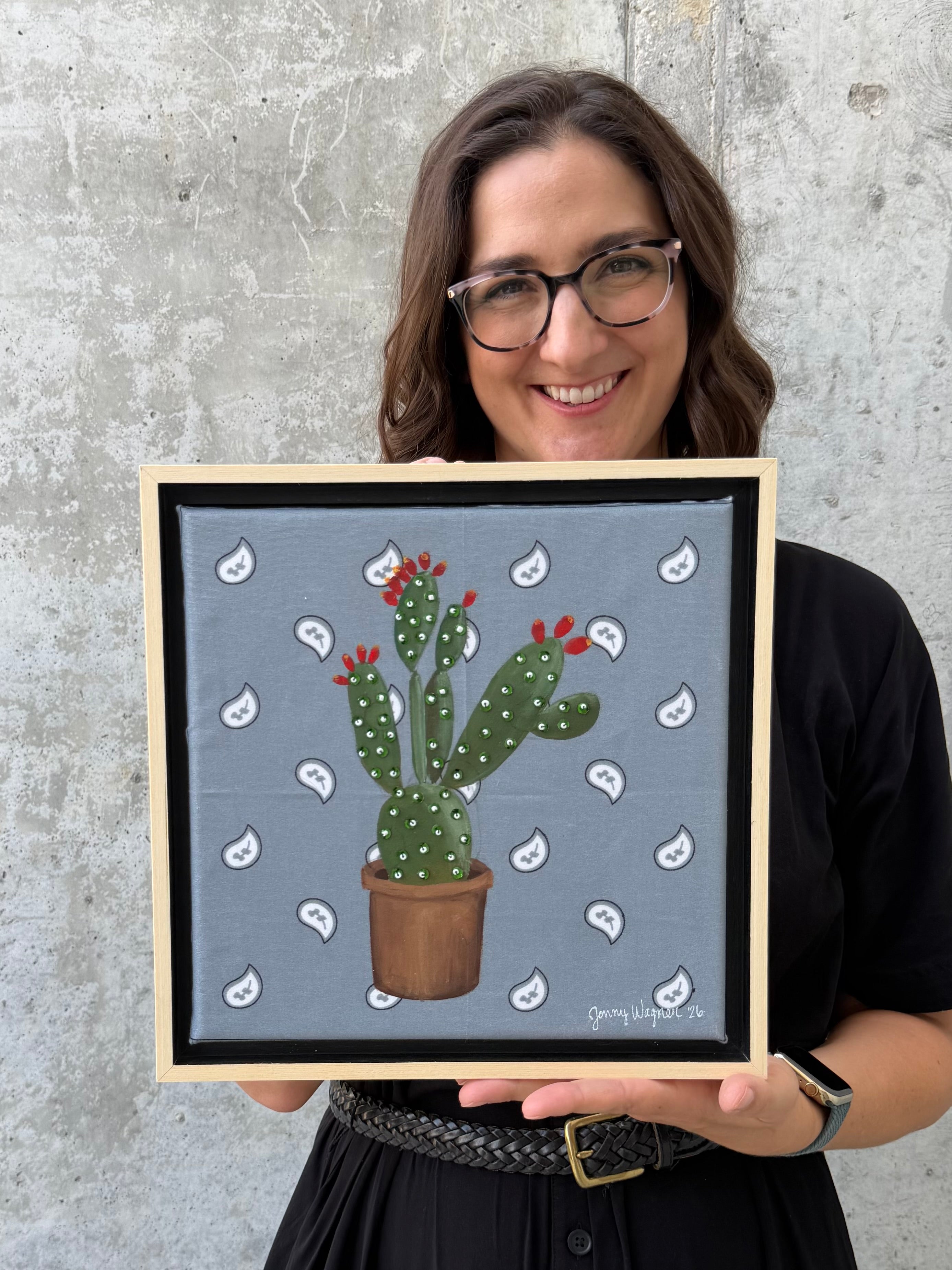 Texas artist Jenny Wagner holding her mixed media original painting, Prickly. This painting features a hand stretched bandana with a potted prickly pear cactus in acrylic paint, with hand beaded glass beads and sequins on top.