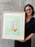 Texas artist, Jenny Wagner, holding her framed original watercolor Bandana Boot. This watercolor shows a light gold boot on top of a light green paisley background with detailed edges. The painting is matted on a floating mount over a white mat and framed in a natural wood frame.