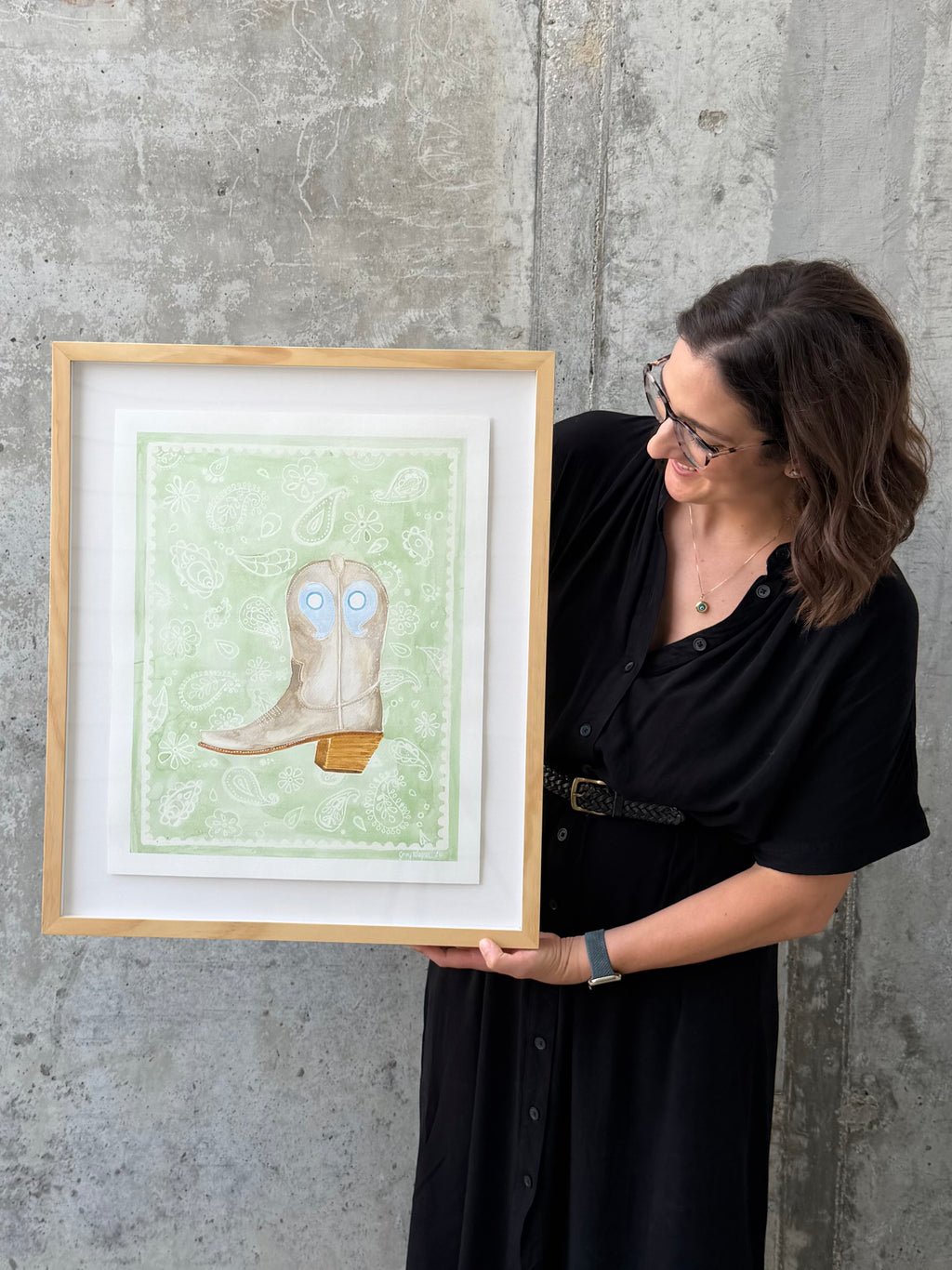 Texas artist, Jenny Wagner, holding her framed original watercolor Bandana Boot. This watercolor shows a light gold boot on top of a light green paisley background with detailed edges. The painting is matted on a floating mount over a white mat and framed in a natural wood frame.