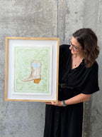 Texas artist, Jenny Wagner, holding her framed original watercolor Bandana Boot. This watercolor shows a light gold boot on top of a light green paisley background with detailed edges. The painting is matted on a floating mount over a white mat and framed in a natural wood frame.