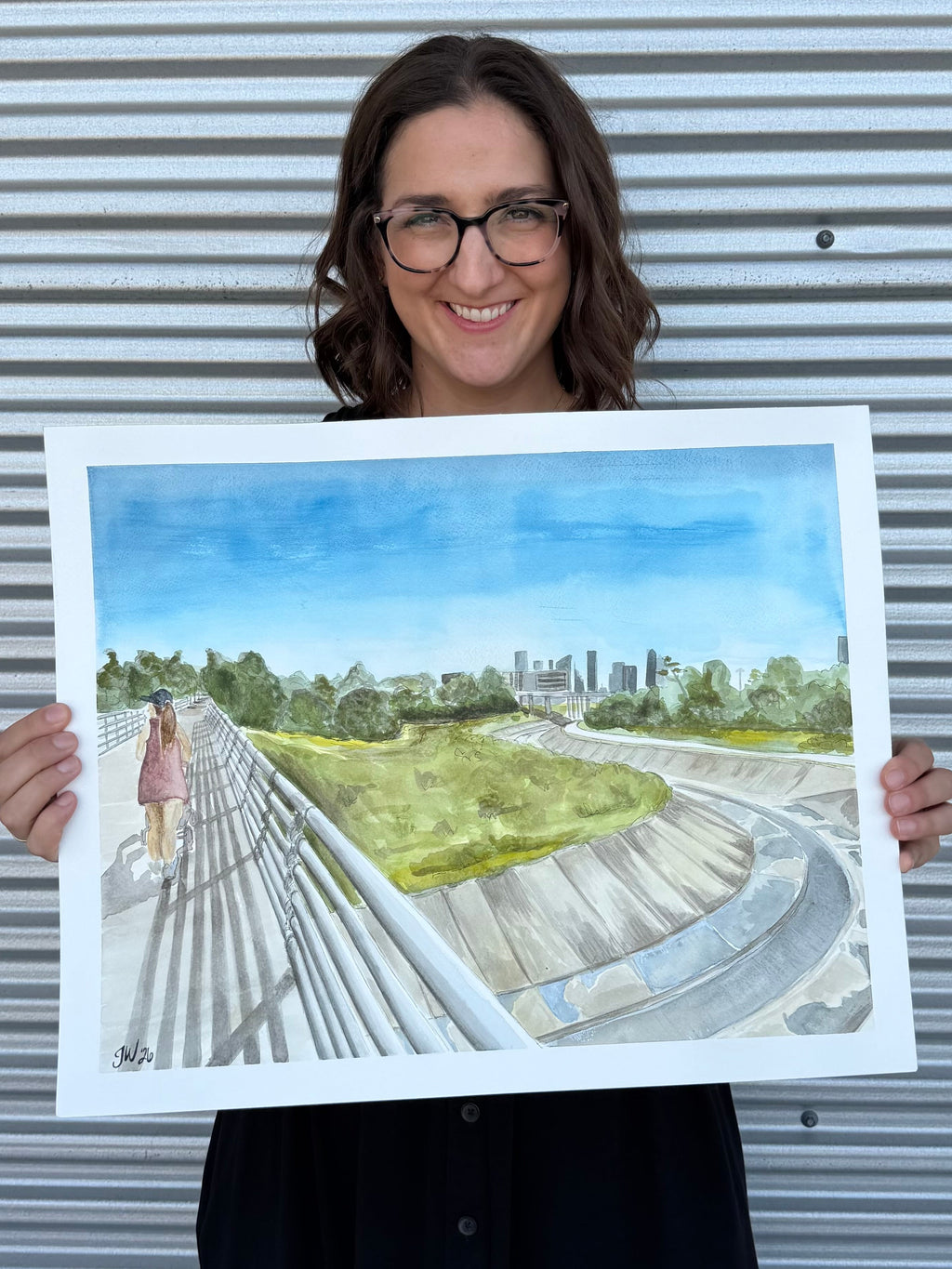 Jenny Wagner holding the original watercolor painting "The Bayou Walk," featuring White Oak Bayou and the Houston Skyline