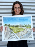 Jenny Wagner holding the original watercolor painting "The Bayou Walk," featuring White Oak Bayou and the Houston Skyline