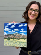 Jenny Wagner holding the original acrylic landscape painting "Colorado to Utah" featuring mountains, blue skies, and summer fields.