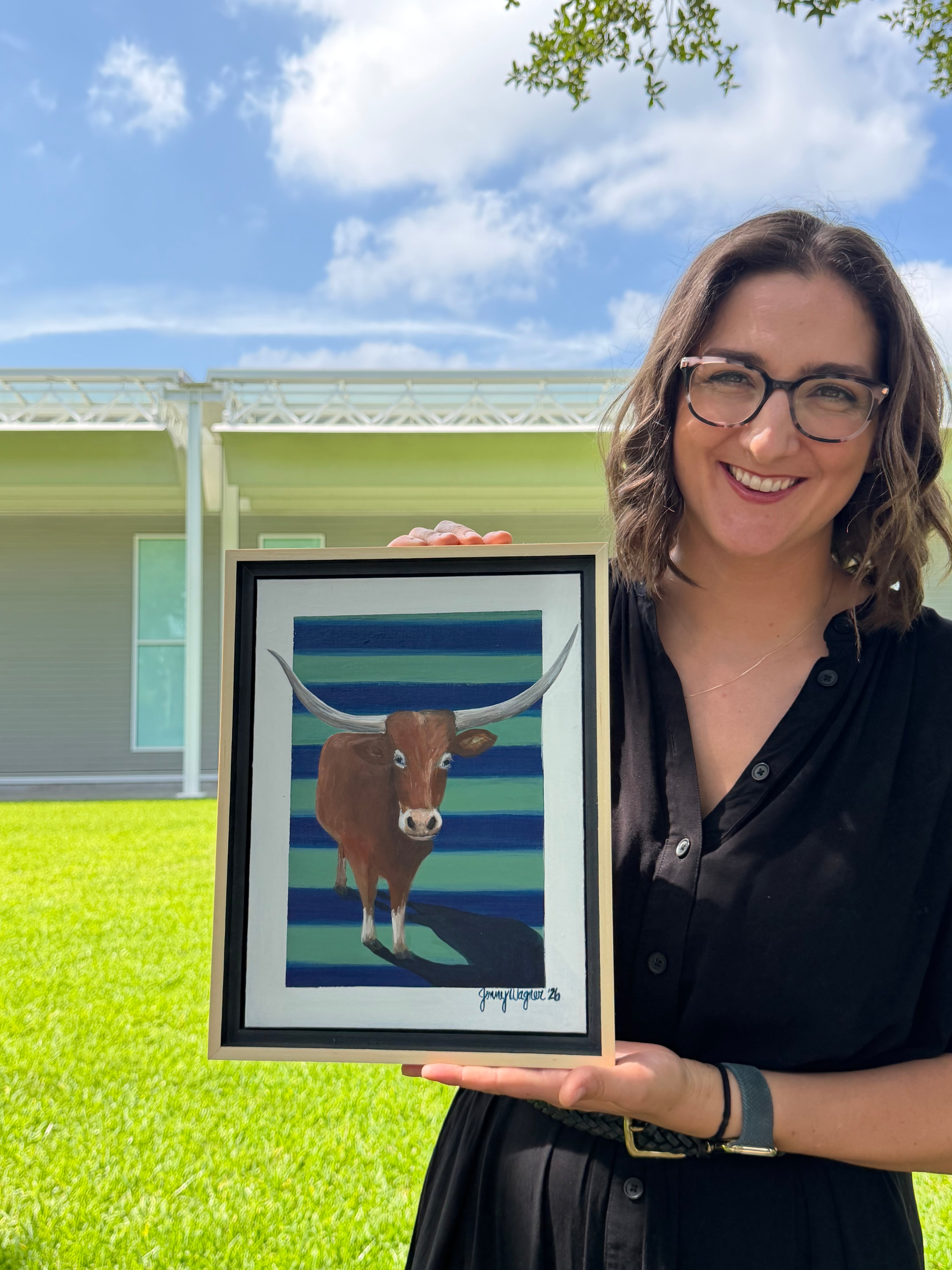 Texas artist, Jenny Wagner shown outdoors in Houston holding her original 9x12 acrylic painting, Ladybird. The painting depicts a brown longhorn on a stripped background with her horns extending through the pattern.