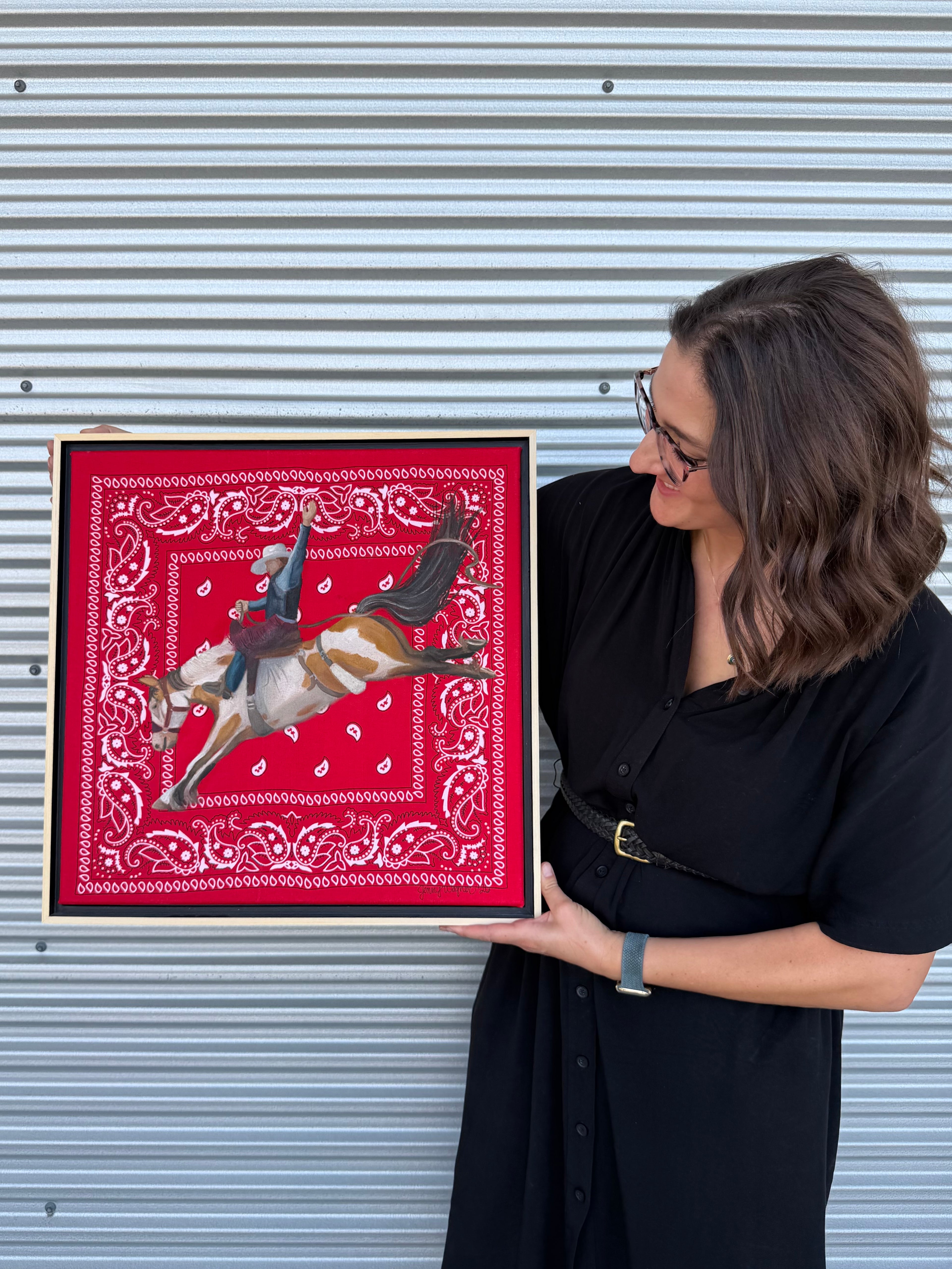 Texas artist, Jenny Wagner, shown holding her original oil painting Hold On. This is the first painting in her ongoing Bandana series. A bronc rider is painted in mid buck atop a hand stretched red bandana.