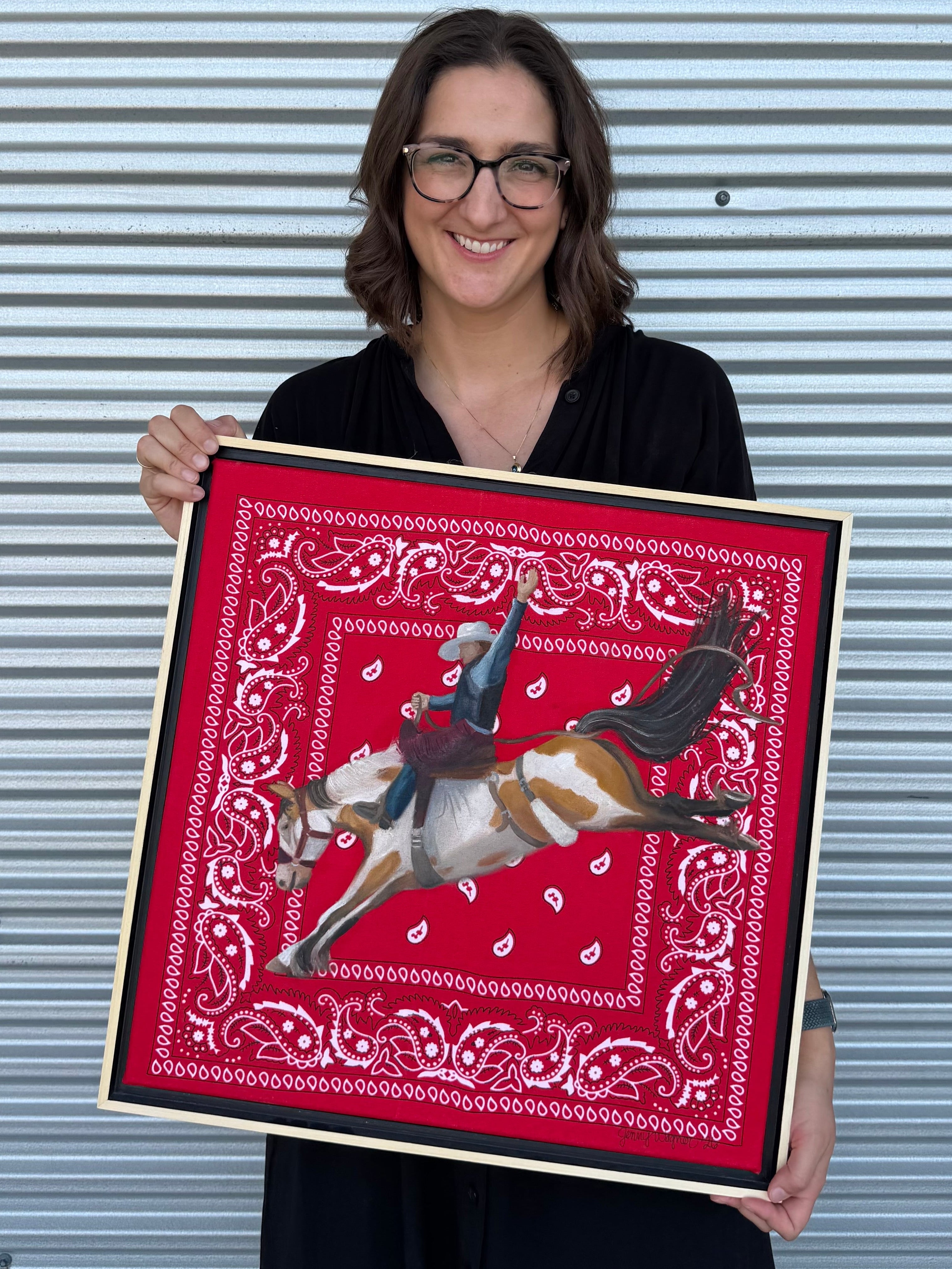 Texas artist, Jenny Wagner, shown holding her original oil painting Hold On. This is the first painting in her ongoing Bandana series. A bronc rider is painted in mid buck atop a hand stretched red bandana.