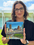 Jenny Wagner holding the original oil painting "Layers," featuring the Houston skyline and Buffalo Bayou
