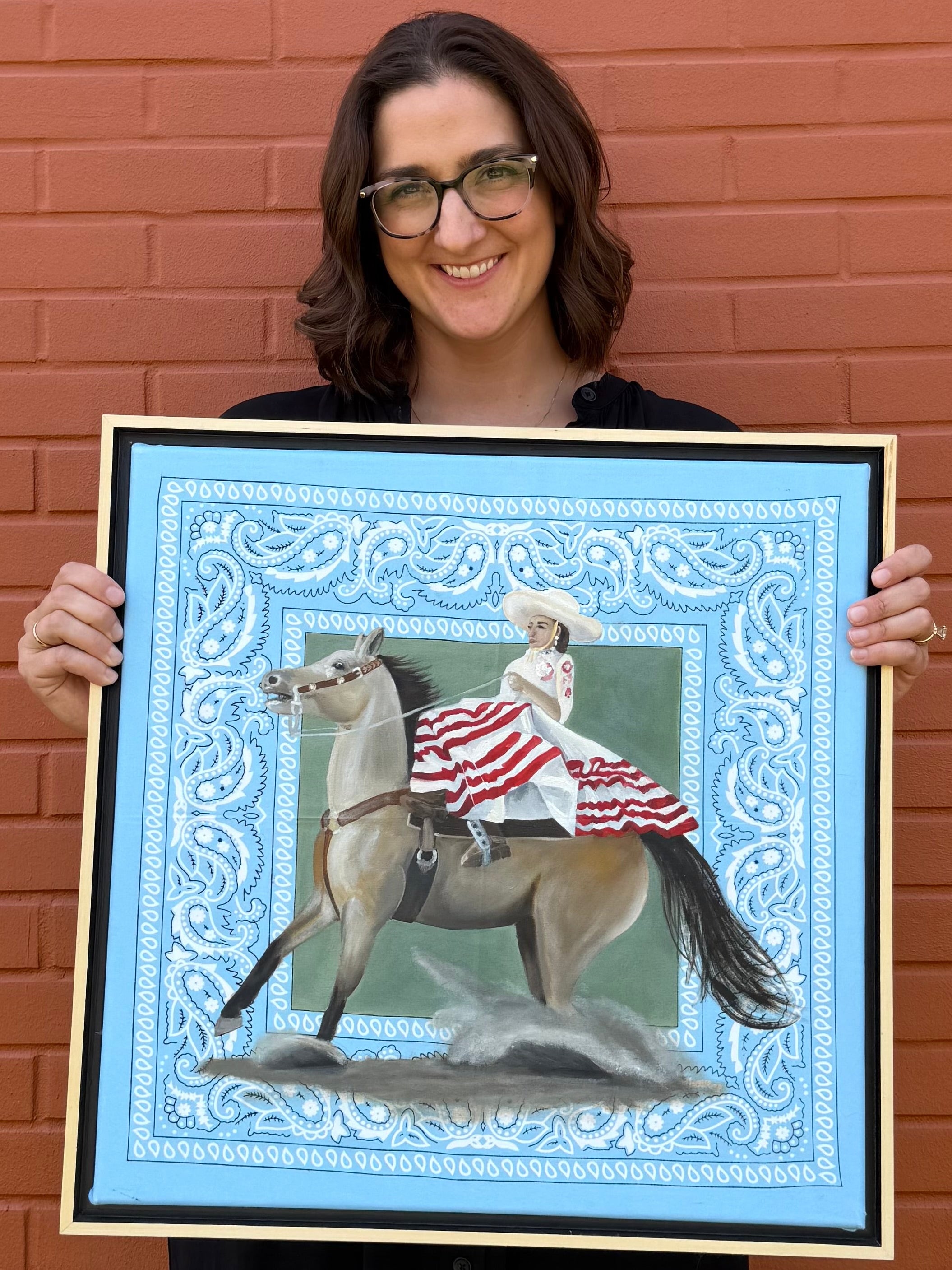 Close up of artist, Jenny Wagner, with her original oil painting La Escaramuza. A Escaramuza Charra rider is shown in traditional dress astride a horse with dirt flying as she comes to a stop. The painting is done directly on a hand stretched pale blue bandana.