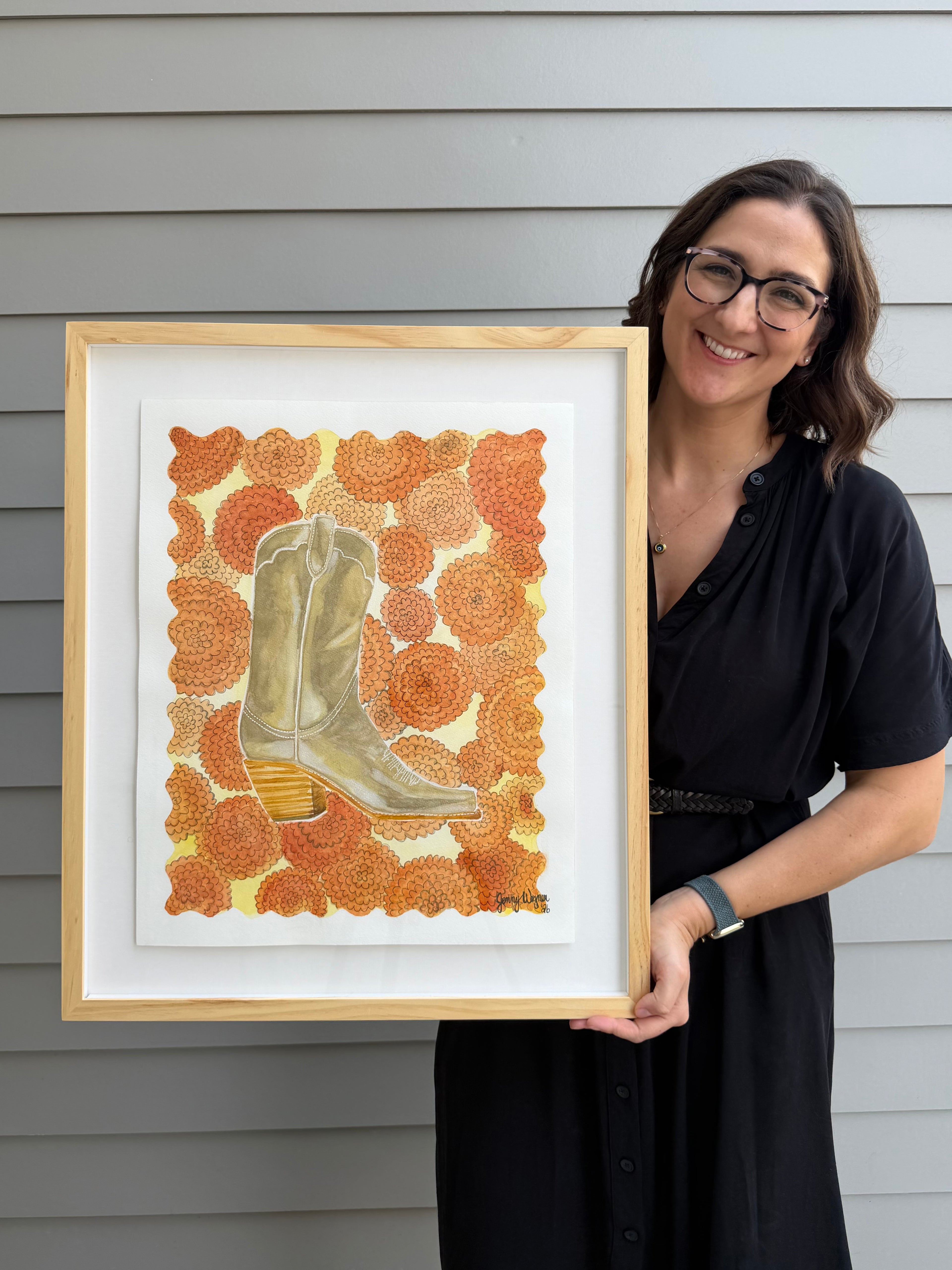 Texas artist, Jenny Wagner holding a framed watercolor original from her boot collection, Standing in Marigolds. This painting is of a metallic gold boot in a bed of orange marigold flowers with a scalloped edge. Matted with a floating mount and framed with a natural wood frame.
