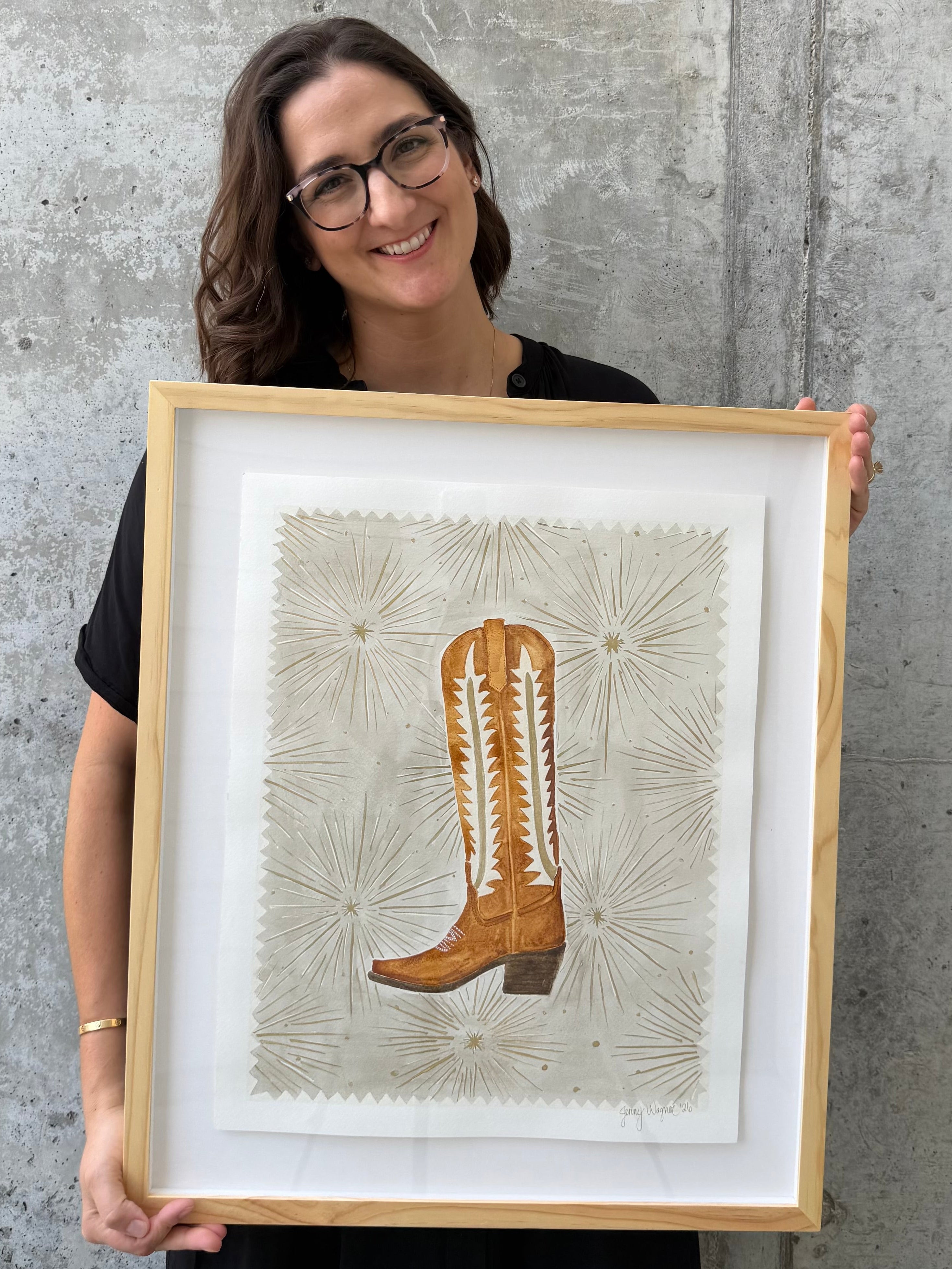 Texas artist Jenny Wagner shown holding her original watercolor "Starburst Boot" featuring a brown cowgirl boot with metallic details. Framed in a natural wood frame.