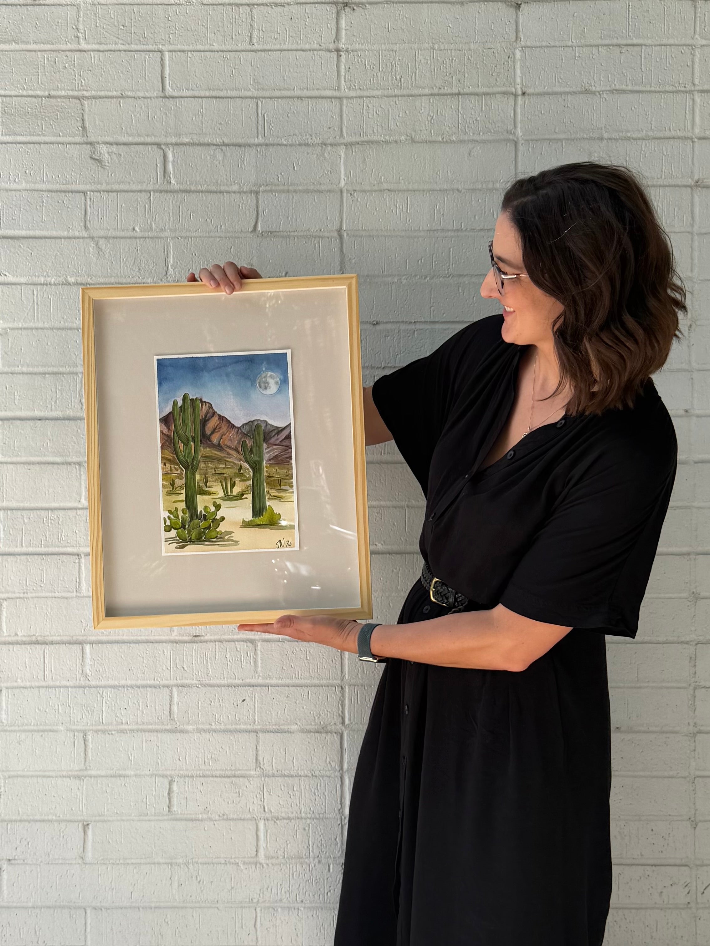 Texas artist, Jenny Wagner, is shown holding her framed original western landscape watercolor, Desert Moon. A desert scene is shown in the evening with a full moon shining over the saguaro cacti.