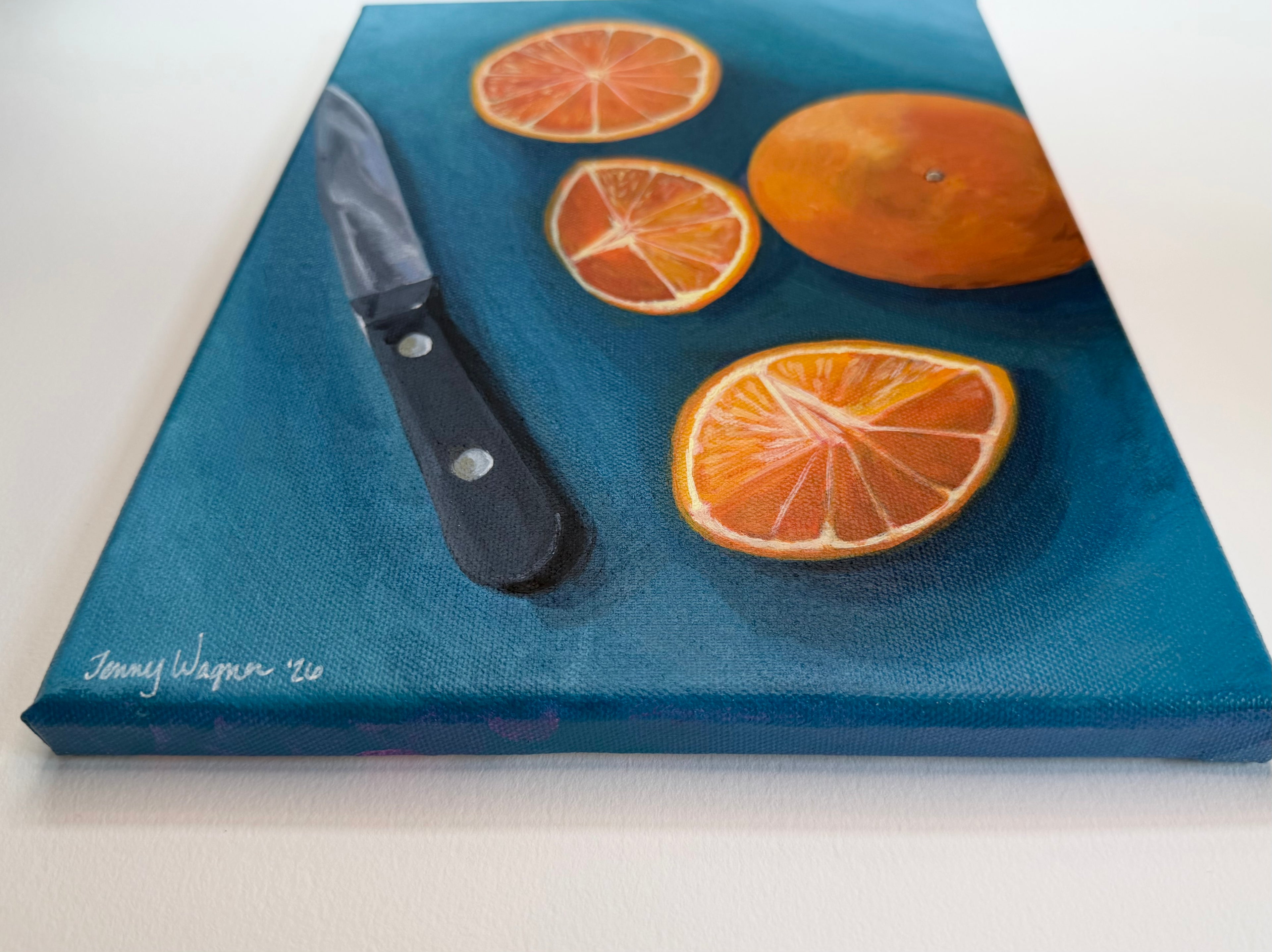 Bottom view of the original 9x12 acrylic painting, Oranges on Blue by Houston artist Jenny Wagner. Signature is shown in bottom left side, and the still life depicts orange slices and a whole orange on a dark blue surface next to a knife, shown from an angled top-down view on a white background.