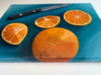 Side view of original 9x12 acrylic painting by Houston artist Jenny Wagner studio. This still life plays with complimentary colors and shows a whole orange and three slices along with the knife that cut the oranges on a blue background.