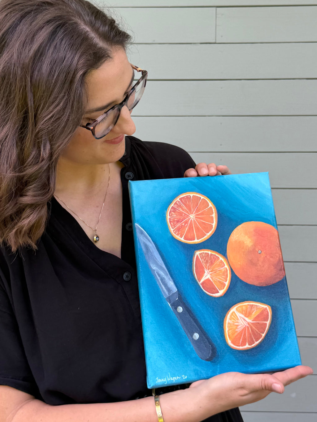 Side view artist Jenny Wagner holding her original 9x12 acrylic painting "Oranges on Blue". This still life plays with complimentary colors and shows a whole orange and three slices along with the knife that cut the oranges on a blue background.