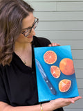 Side view artist Jenny Wagner holding her original 9x12 acrylic painting "Oranges on Blue". This still life plays with complimentary colors and shows a whole orange and three slices along with the knife that cut the oranges on a blue background.