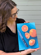 Side view artist Jenny Wagner holding her original 9x12 acrylic painting "Oranges on Blue". This still life plays with complimentary colors and shows a whole orange and three slices along with the knife that cut the oranges on a blue background.